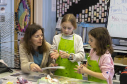 Woman and two children do a paper art activity at a table. The children are wearing bright green aprons.