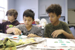 Three boys on a field trip are seated at a table in WMA's Studio. They are stamping papers with colorful inks.