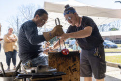 Two men hold large metal tongs together while shaping hot glass over a wooden bench.