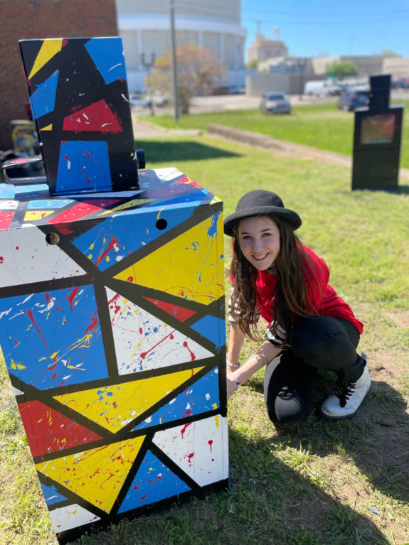 A teenager is painting a newspaper box with bright, primary colors.
