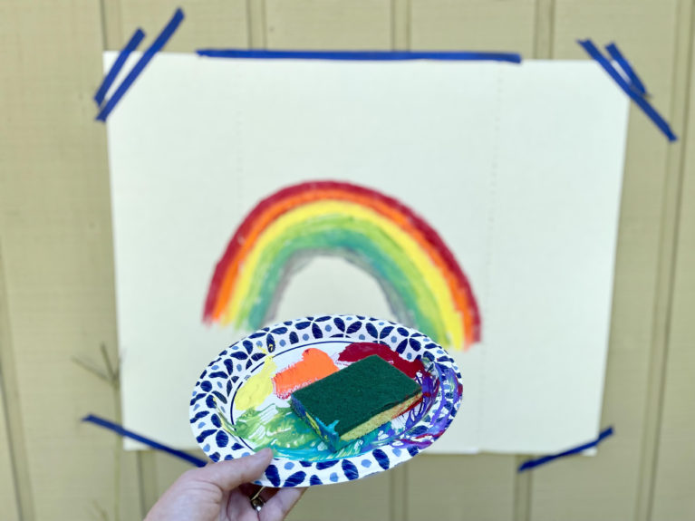 A sponge painted rainbow on carboard that has been taped to the side of a house.