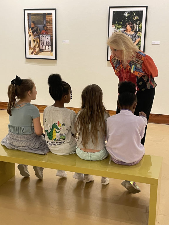 Four young students sit on a bench while their docent discusses the artwork in front of them.