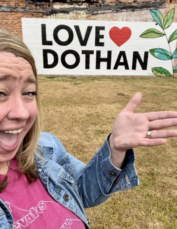 A woman happily stands in front of a mural that reads, "Love Dothan."