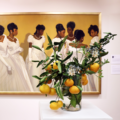 A floral display of greenery, satsumas, and white flowers sits on a white pedestal in the foreground. A large, horizontal painting of Dale Kennington's "The Debutantes" is in the background.