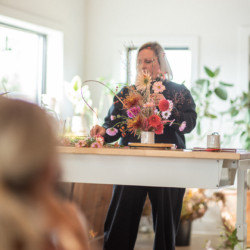 A woman (Holly Carlisle) is standing behind a white, elevated work table. She is arranging pink, mauve, and orange flowers in a white container on the table. 