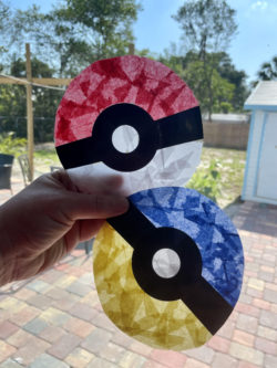 A hand holds two Pokeball suncatchers made with tissue paper and contact paper. They are held in front of a window and one is white and red and the other is yellow and blue. 