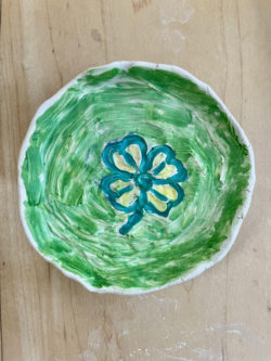 A green handmade clay trinket dish has a green and yellow four leaf cover design in the middle. The dish lays on a light wood table. 